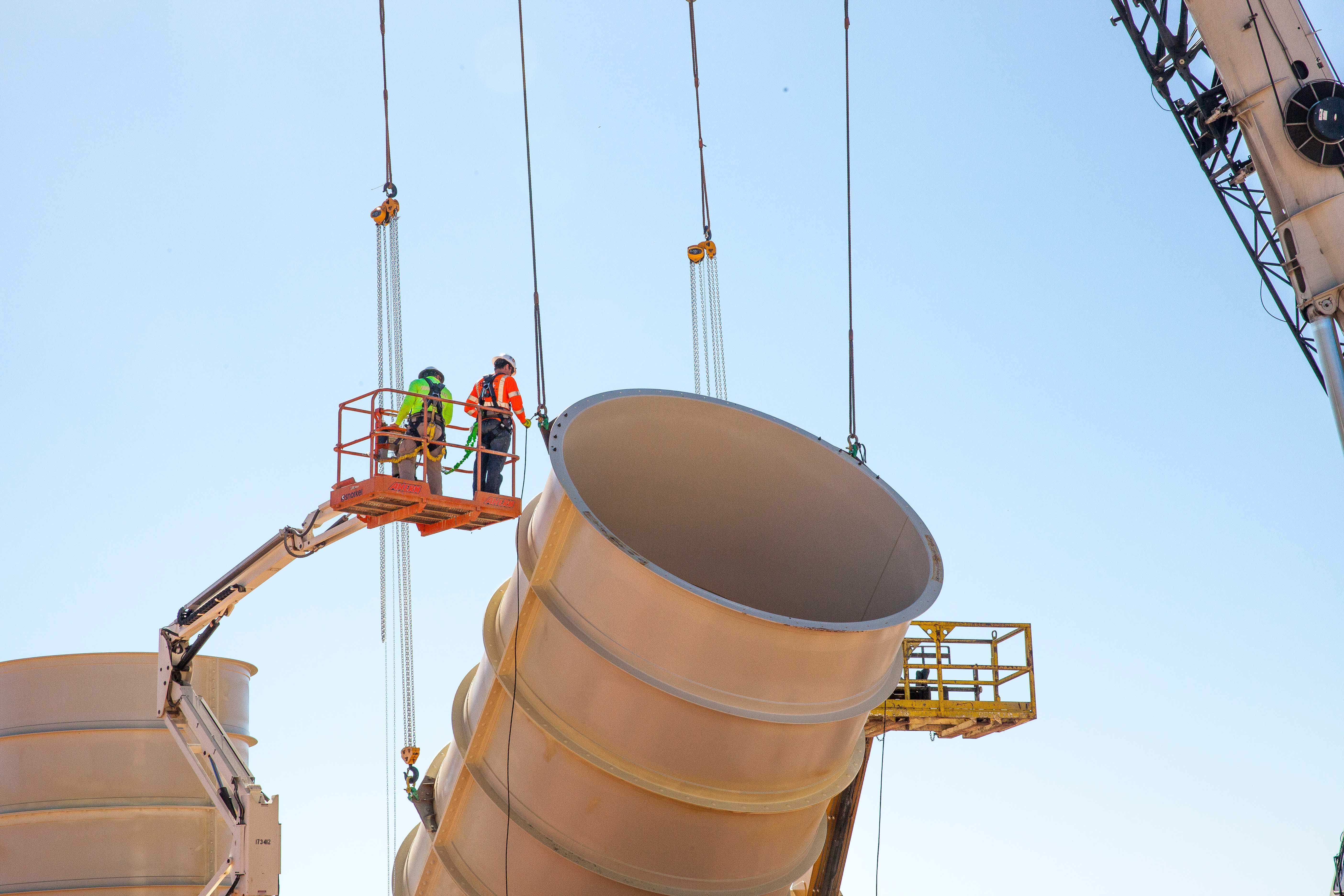 Riggers guiding metal ductwork as it's lifted into a trench at the new utility shaft